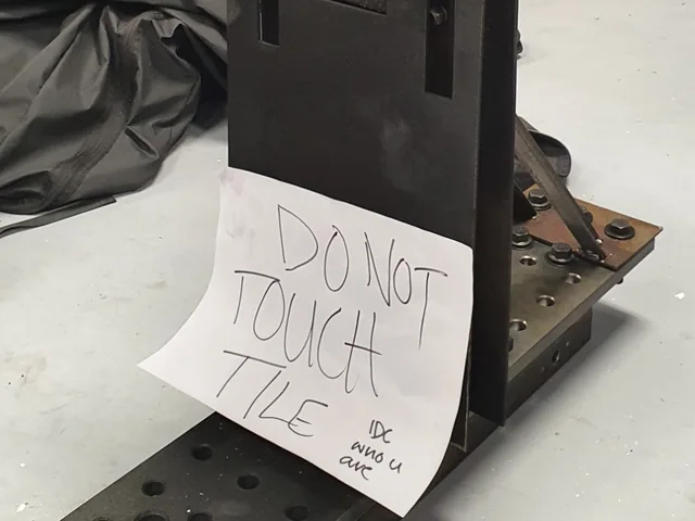The finished rocket firing stand: a black steel plate on a bolted base rail, with a handwritten sign reading &ldquo;Do not touch tile, IDC who u are&rdquo;.