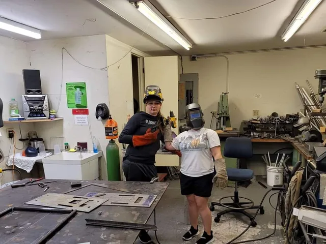 In the welding shop, me with my helmet up next to a student in a welding helmet and gloves, steel plates for the stand laid out on the table.