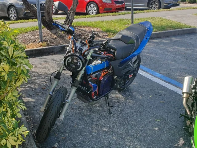 The motorcycle running without its tank: the blue battery module, controller and wiring exposed in the frame behind a round LED headlight.