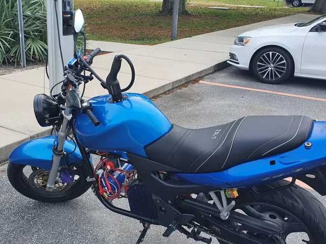The motorcycle plugged into a public J1772 charging station, the cable running into the old fuel filler.