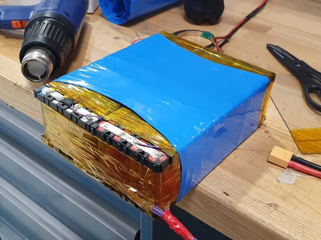 A finished battery module wrapped in amber insulating tape and blue heat-shrink, with the heat gun and balance leads beside it.