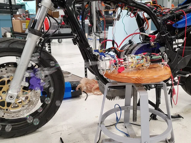 A clear acrylic bubble machine on a stool next to the motorcycle, bubbles floating past the front wheel.