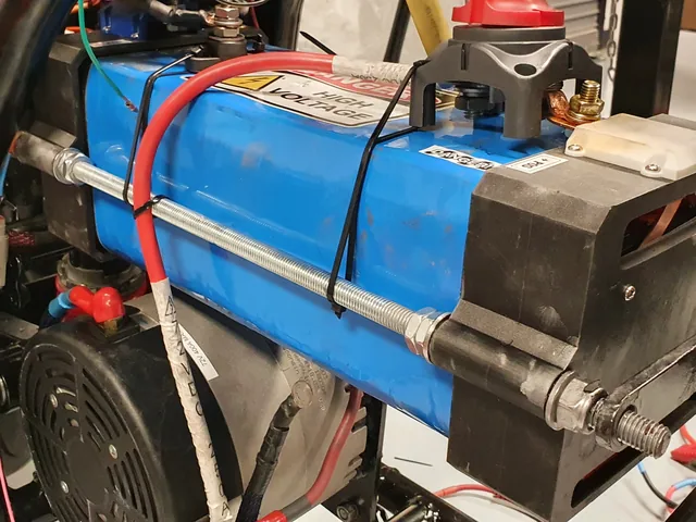 The blue battery pack held between black end plates by threaded rods, mounted in the motorcycle frame and marked Danger High Voltage.