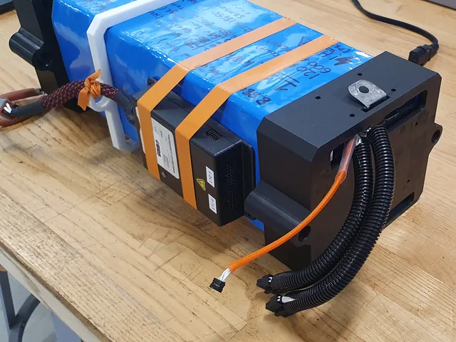 A long battery wrapped in blue heat-shrink with orange straps and a black 3D-printed end cap, cables leaving through corrugated conduit.